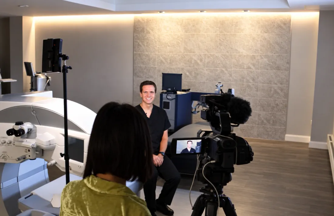 A doctor in scrubs smiling during a filmed interview in a treatment room, seen behind a video camera